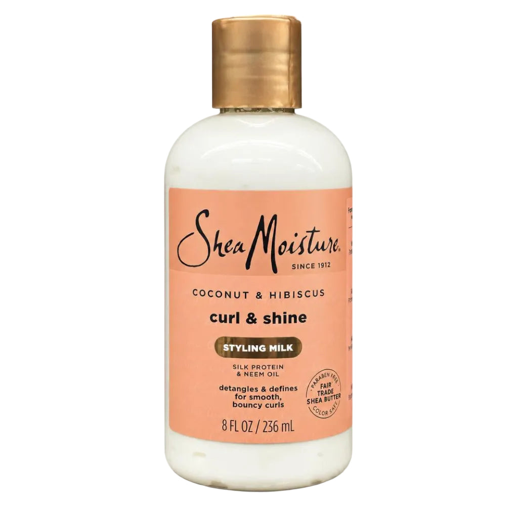 A bottle of SheaMoisture Curl & Shine Styling Milk in Coconut & Hibiscus, featuring an off-white bottle, a peach-colored label, and a metallic gold cap, set against a plain white background.